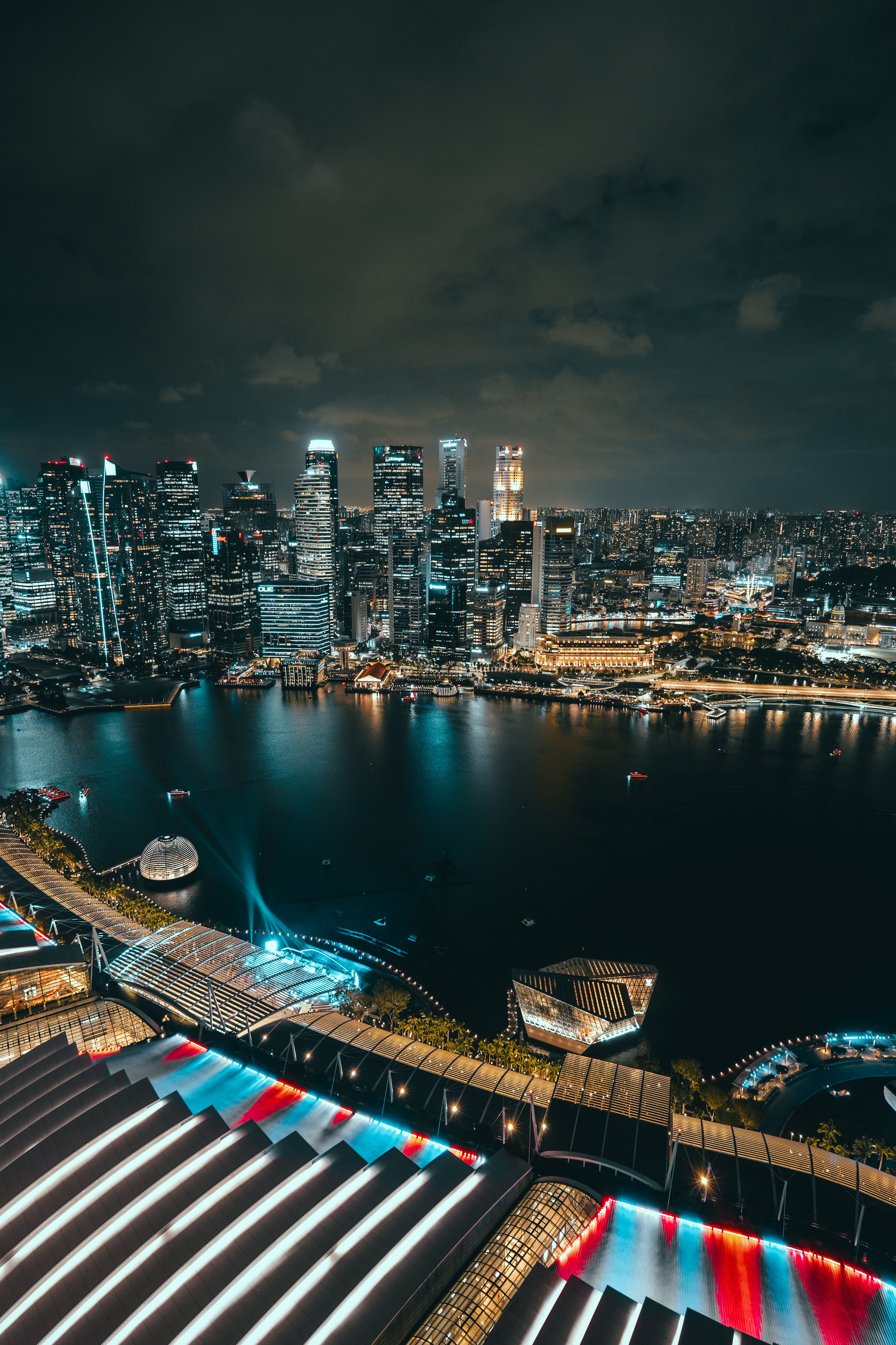 A night-time aerial view across a harbor shows a dense, illuminated city skyline in the distance, a curved lit bridge and an angular glass building along the waterfront, and in the foreground a scalloped rooftop terrace streaked with red, white, and cyan light trails under a dark, cloudy sky.