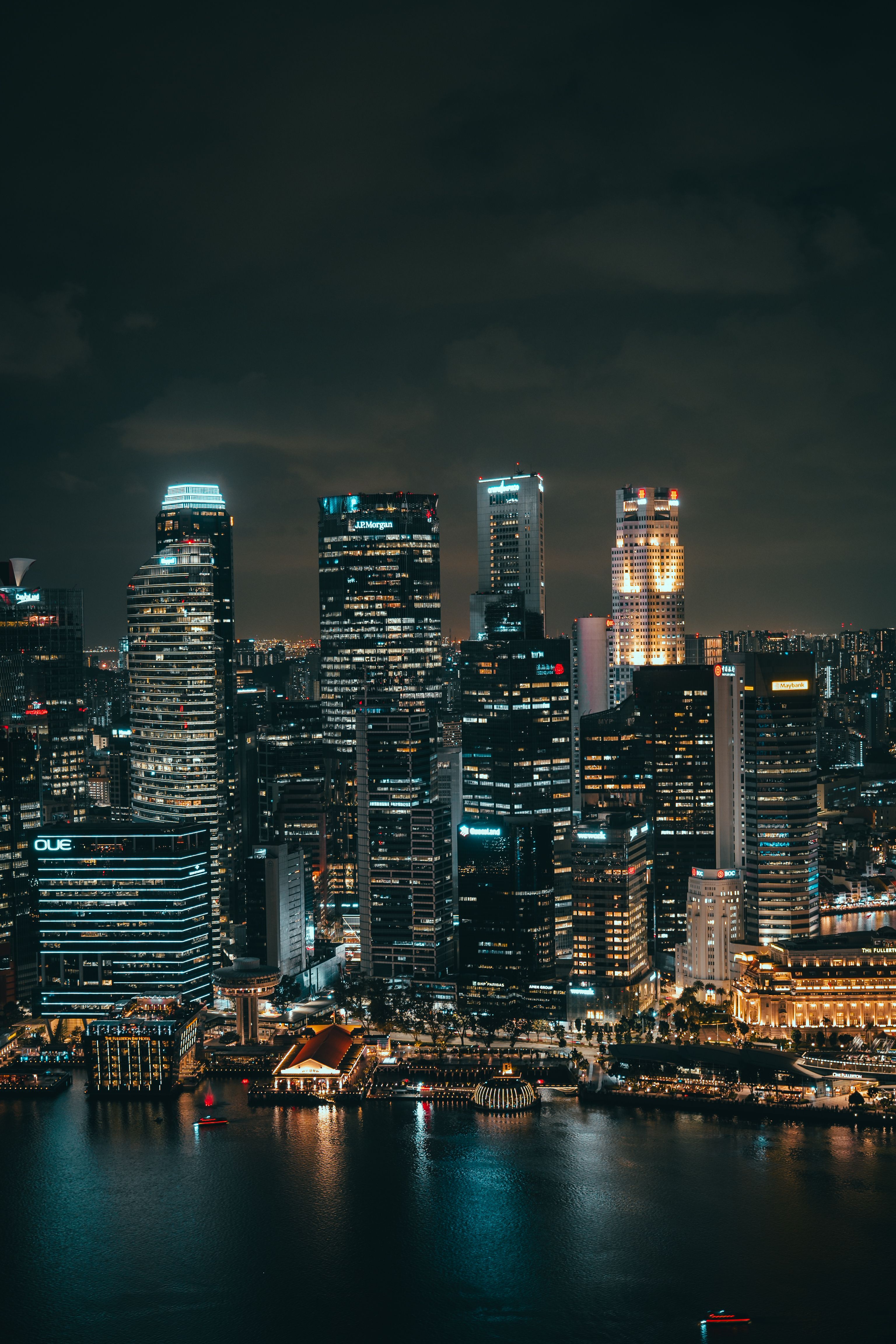 A nighttime skyline of tall illuminated skyscrapers rises above a waterfront, their lights in white, blue, and amber reflected in the dark water below, with a lit domed pavilion and rows of lower buildings along the shoreline.