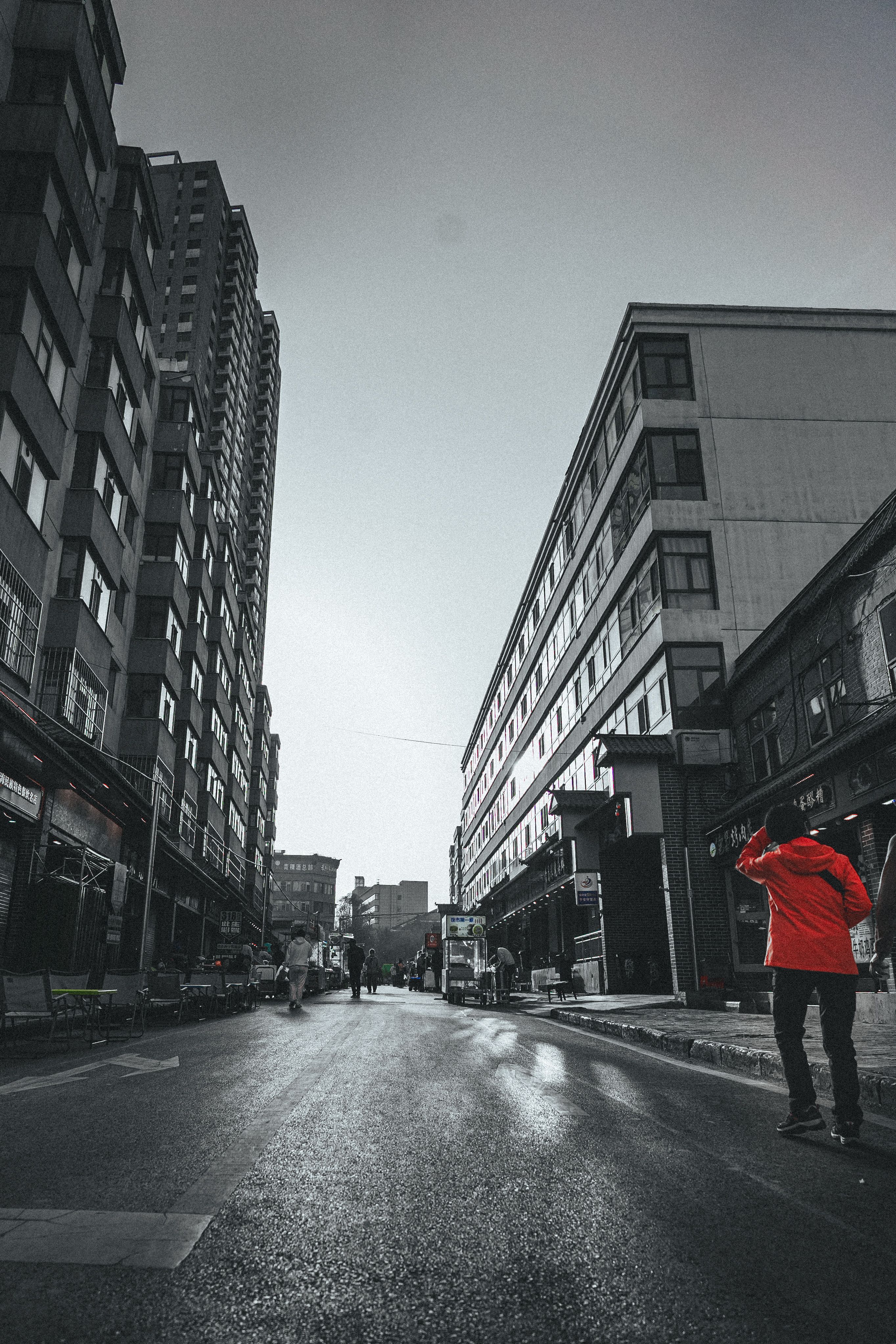 A desaturated street scene of a wet urban road flanked by tall apartment buildings that converge toward a hazy overcast sky, with a person in a red jacket standing in the right foreground, hand raised to their head, and several pedestrians and market carts further down the street.