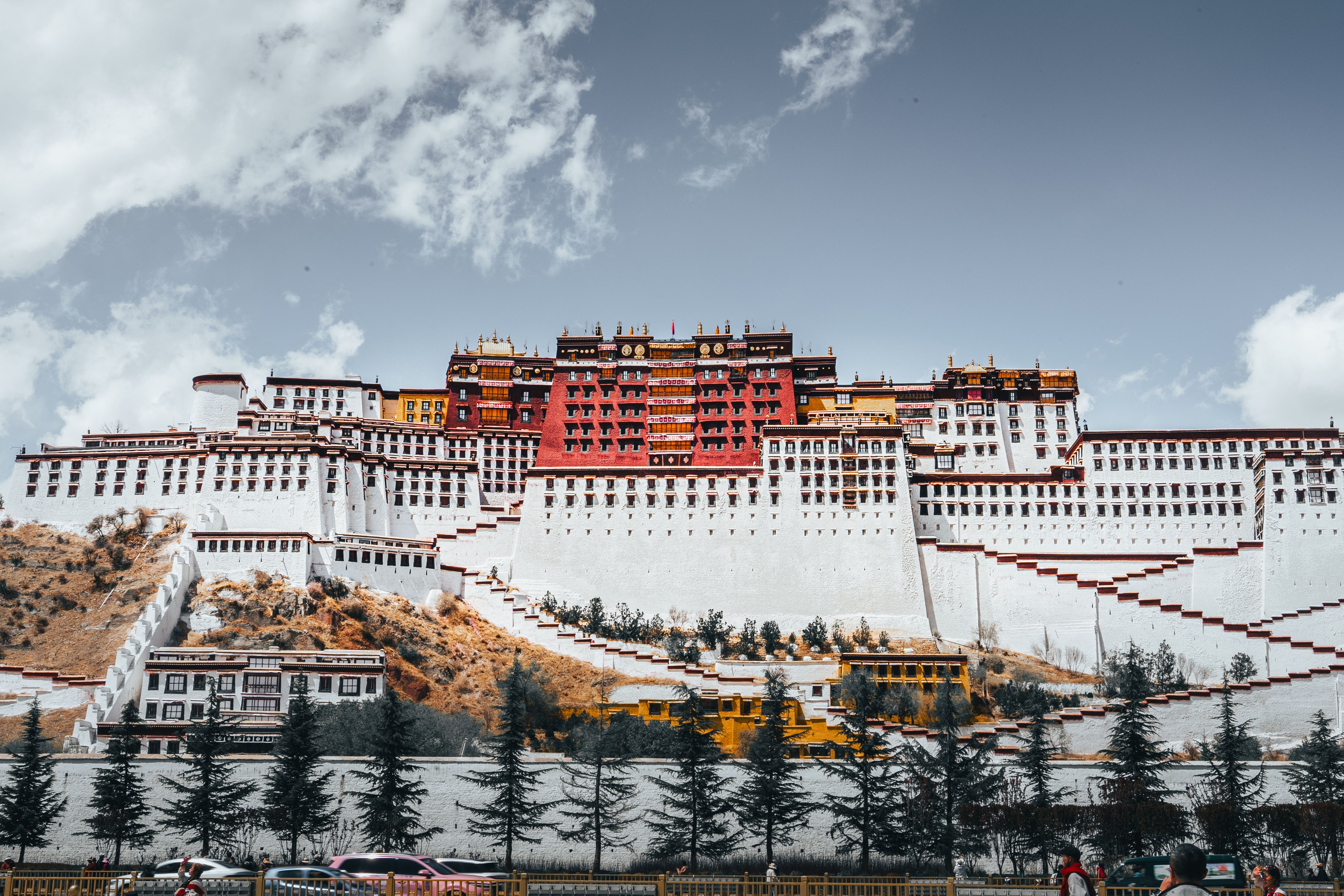 A large white-and-red tiered palace with rows of small square windows and zigzagging stone staircases spans a hillside under a bright, partly cloudy sky, with a line of dark conifer trees, parked cars, and pedestrians along a roadside fence in the foreground.