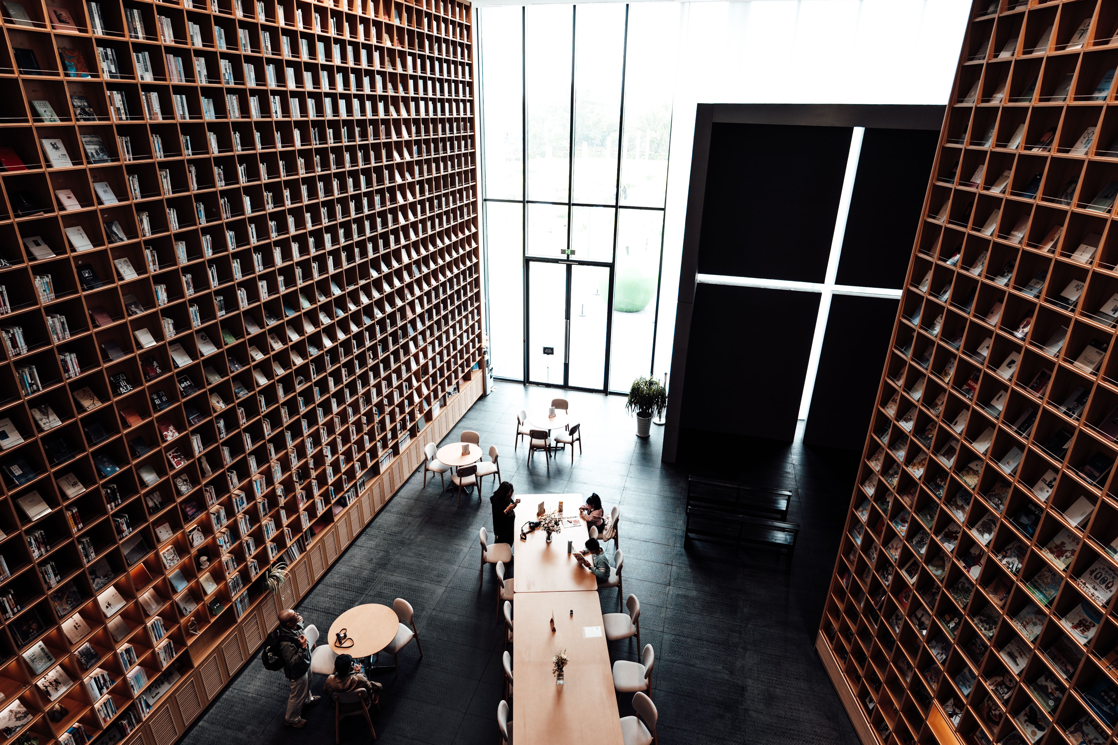 A high-angle interior view of a library with two towering wood-grid bookshelves rising to a high ceiling on either side, a few people seated at tables on the dark floor below, a tall glass entrance at the far end letting in bright daylight, and a dark wall with an illuminated cross-shaped light cutout on the right.