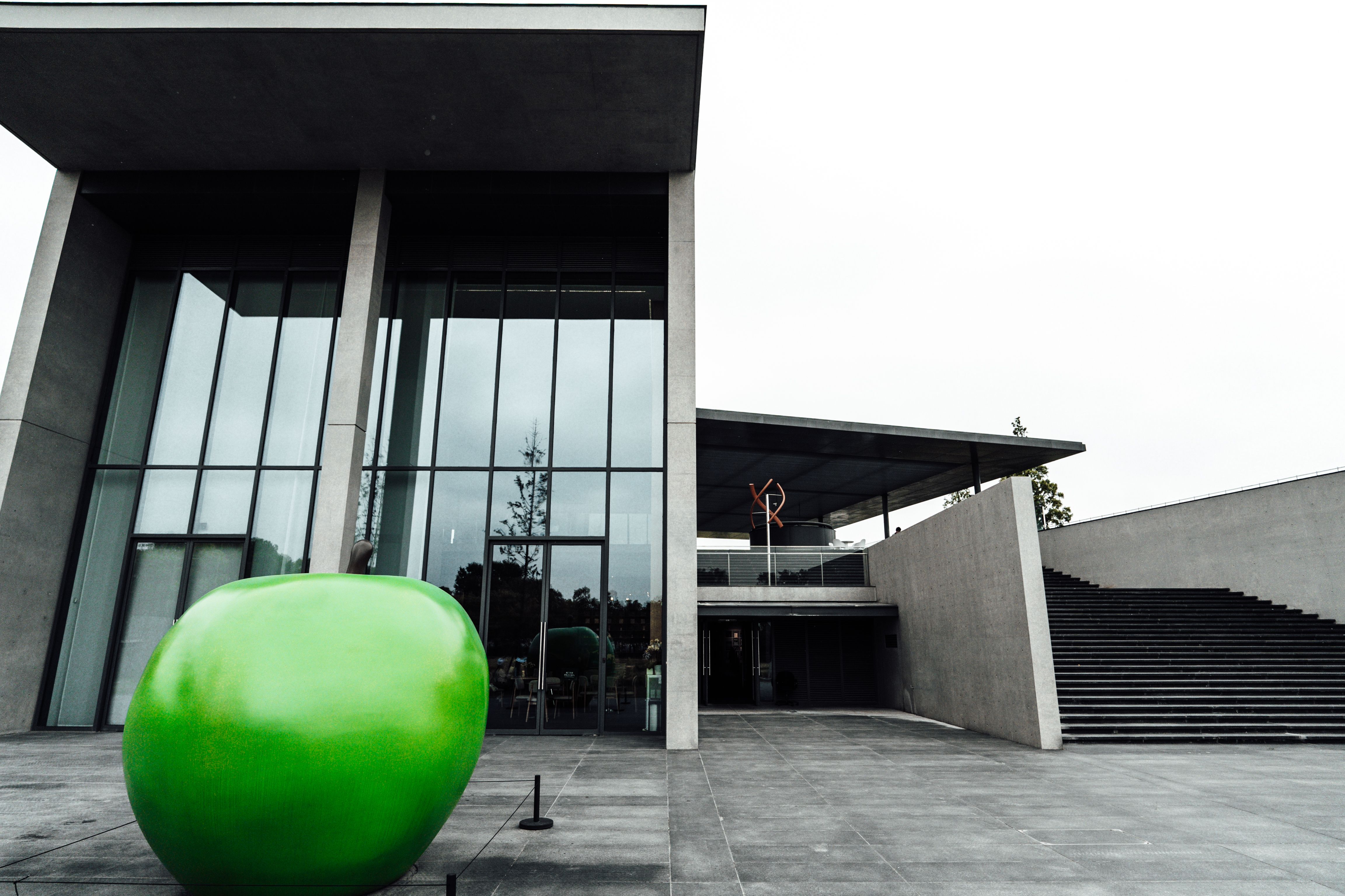 A large green apple sculpture sits on a concrete plaza in front of a modern building with a tall glass curtain-wall facade and a wide outdoor staircase, photographed under overcast light with the rest of the scene rendered in grayscale.