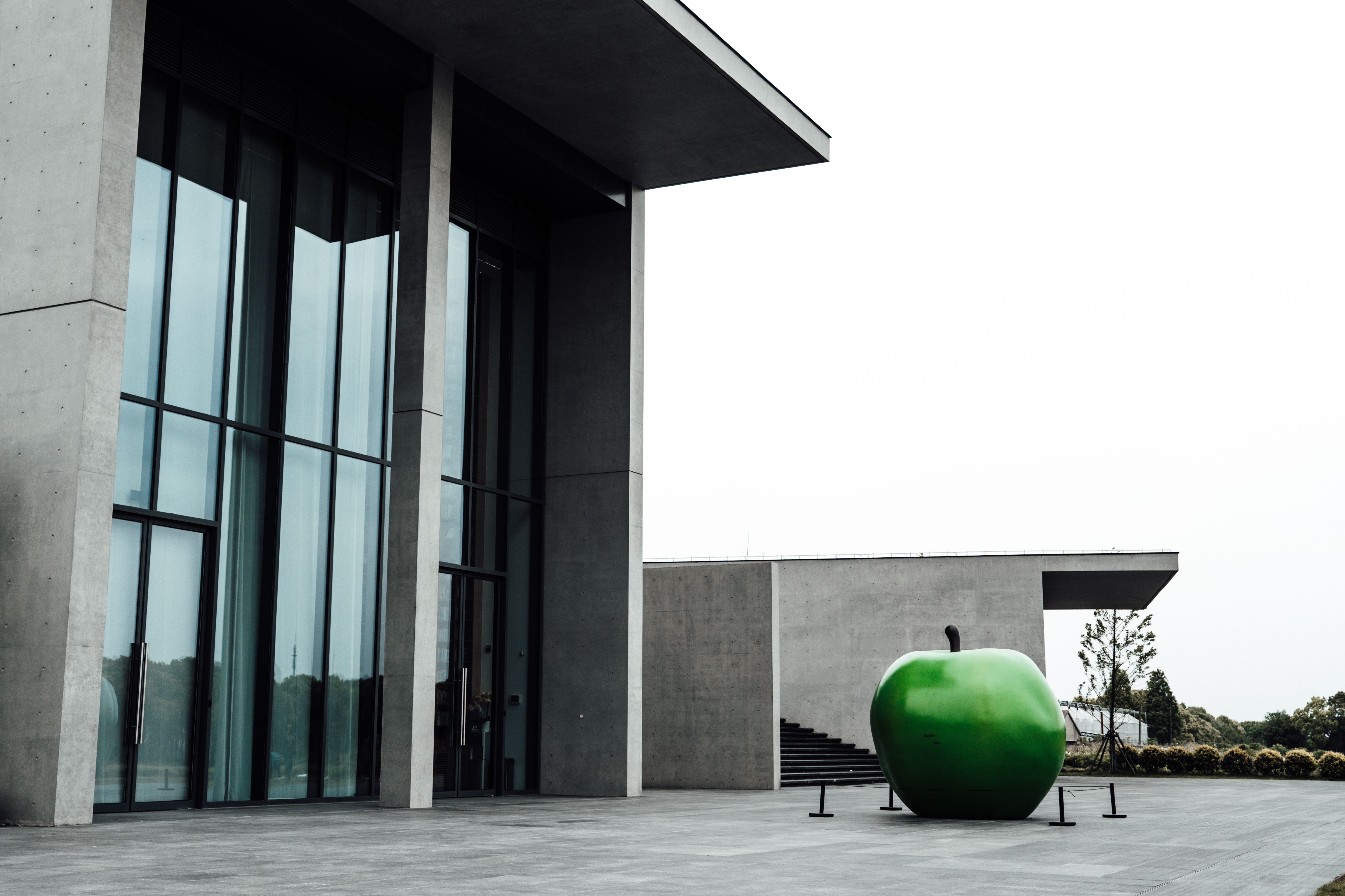 A large green apple sculpture with a black stem stands on a paved concrete plaza beside a minimalist concrete building with tall glass-and-steel windows and a flat cantilevered roof, under a flat white overcast sky with trees visible in the distance.