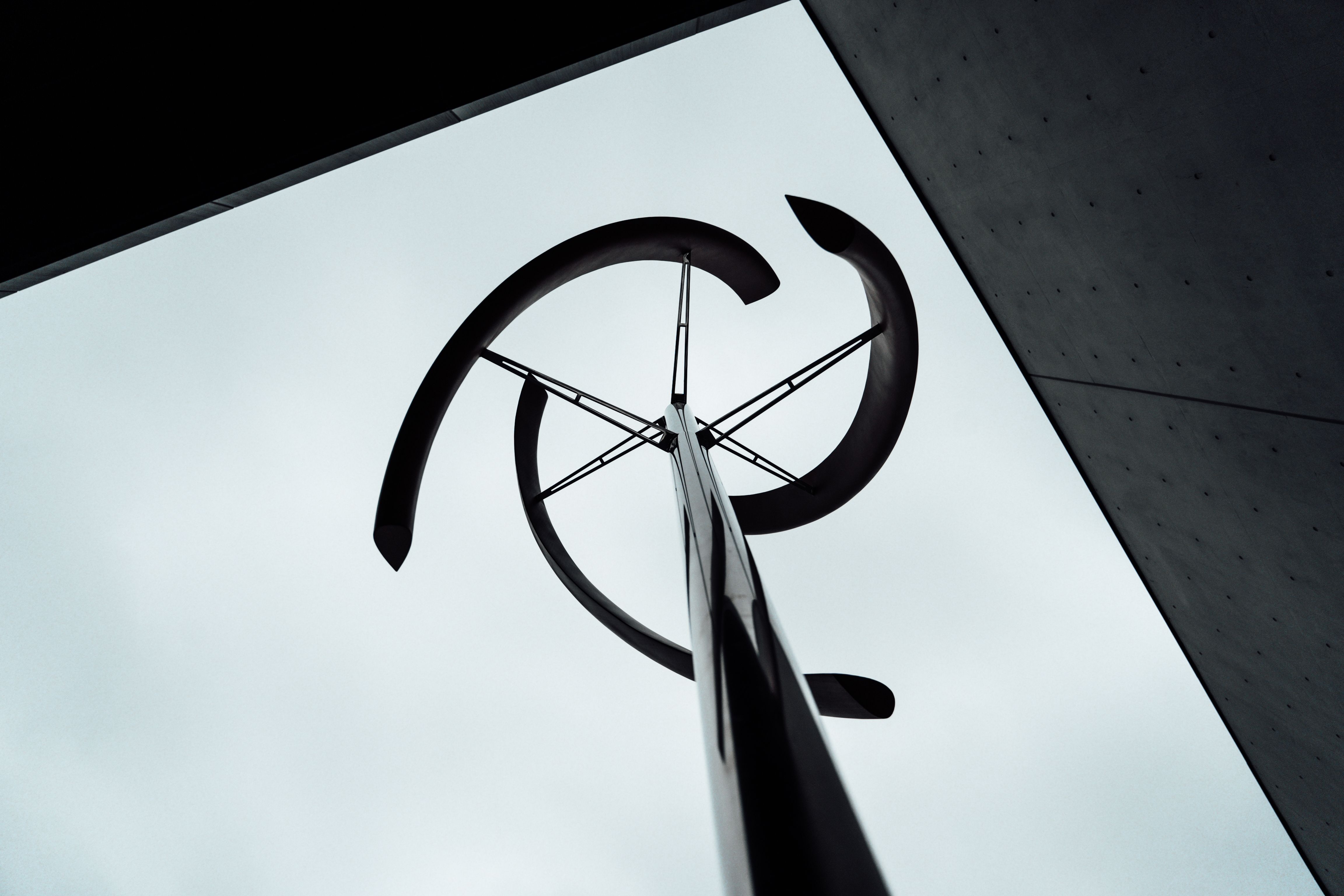 A low-angle photograph looking straight up at a curved, pinwheel-shaped metal sculpture mounted atop a slender pole, silhouetted against an overcast white sky, with the sharp diagonal edge of a dark building facade cutting across the upper right corner of the frame.