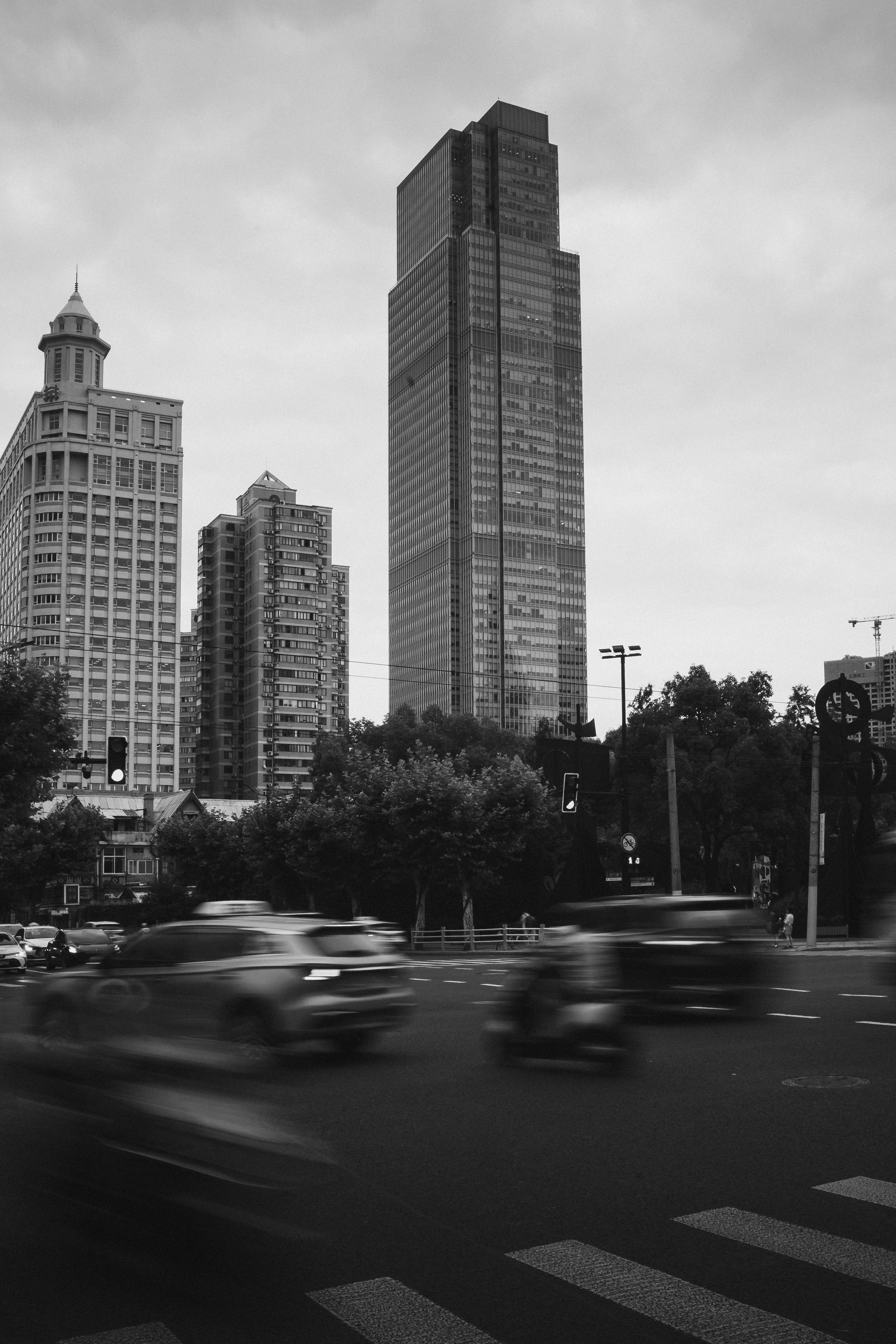 Black-and-white photograph of a city intersection with a tall glass skyscraper rising behind an older high-rise with a domed turret and residential towers, trees lining the roadside, and blurred cars and a motorcycle crossing the street in the foreground under an overcast sky.