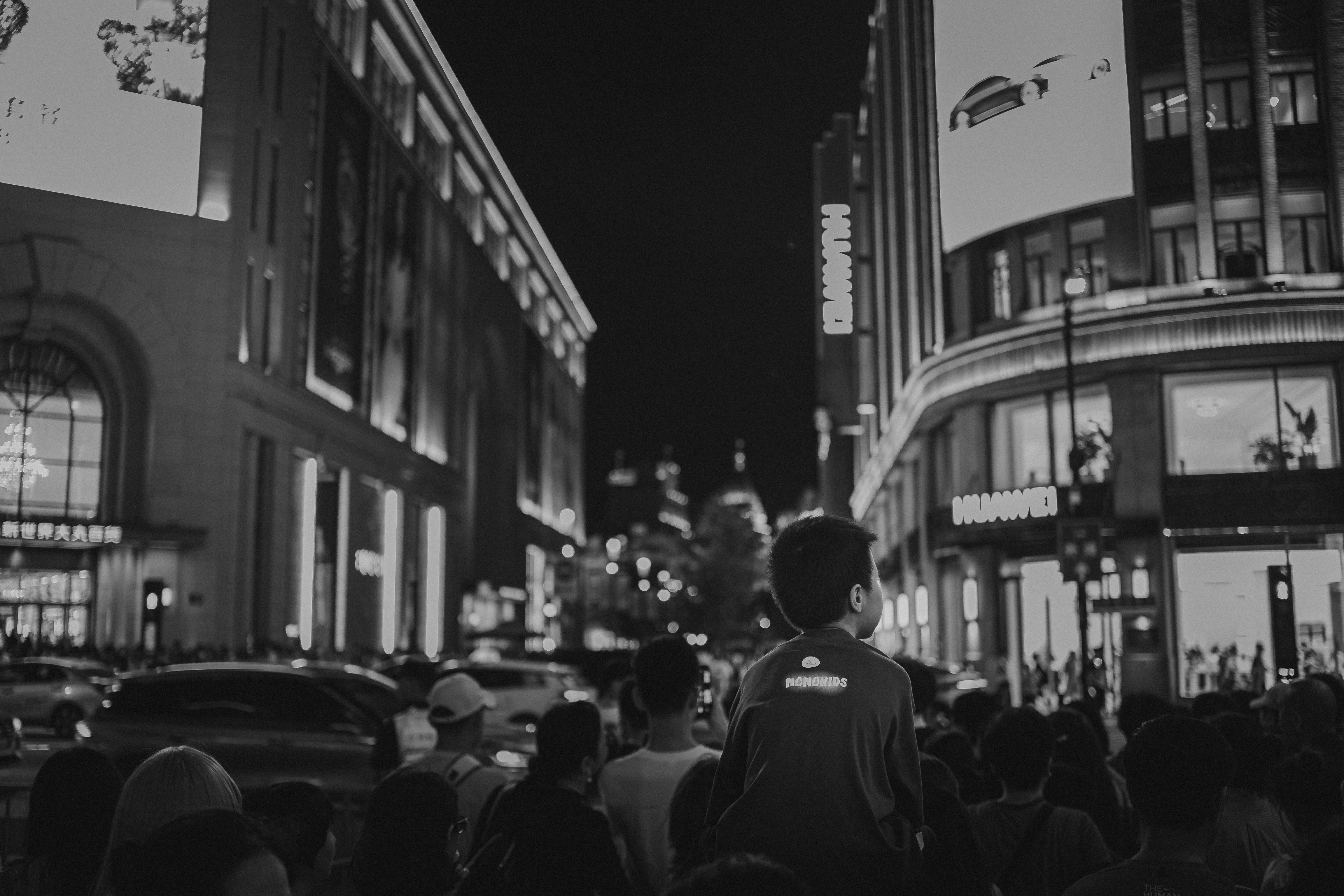 Black-and-white night photograph of a crowded city street, with a child raised above the crowd on an adult's shoulders in the foreground looking up at tall illuminated buildings and a large digital billboard showing an image of a car.