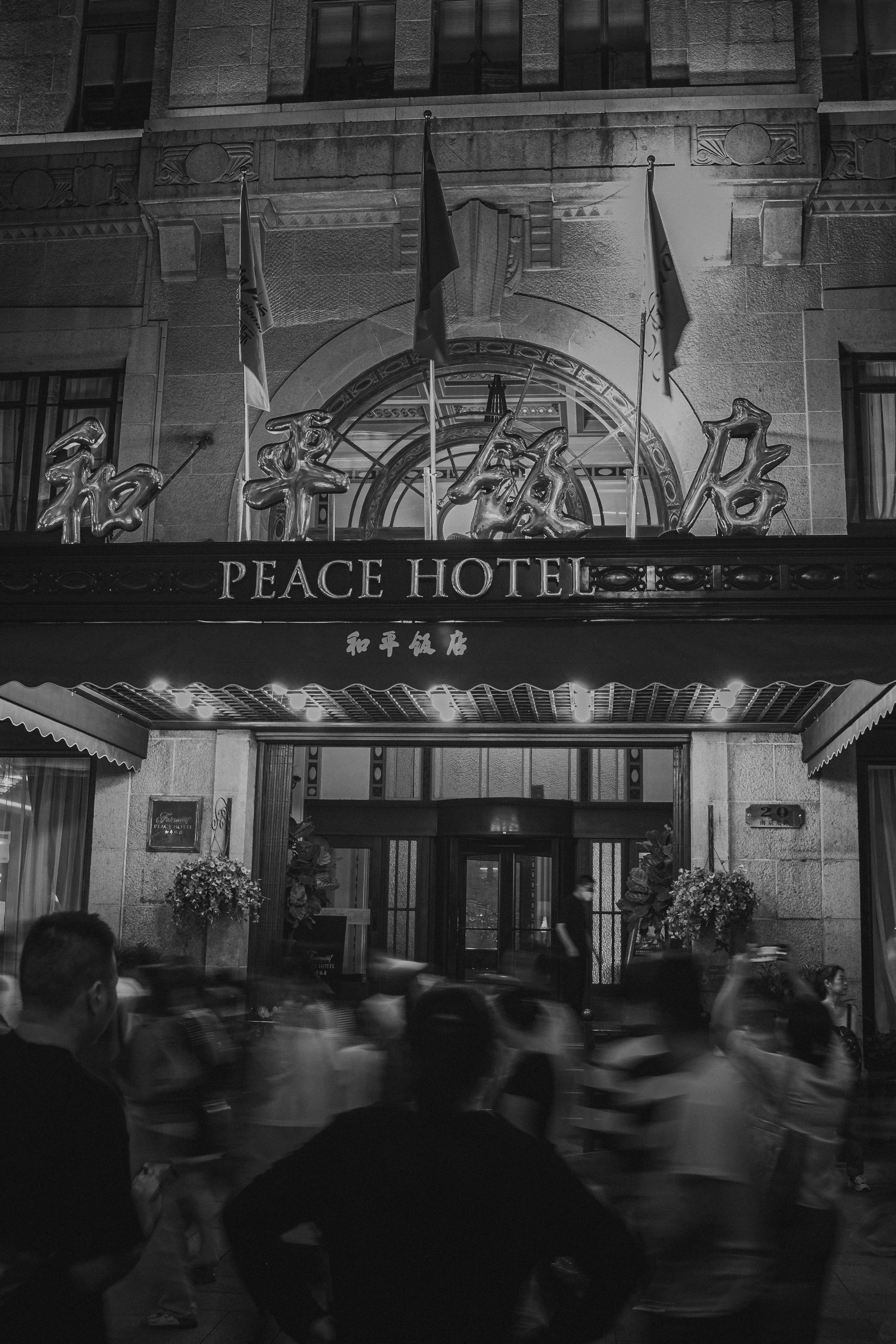 Black-and-white night photograph of an ornate stone hotel facade with three flags and large reflective Chinese characters above a marquee reading "PEACE HOTEL," a light-strung canopy over the entrance, one person standing still near the doorway, and a crowd of people blurred by motion in the foreground.