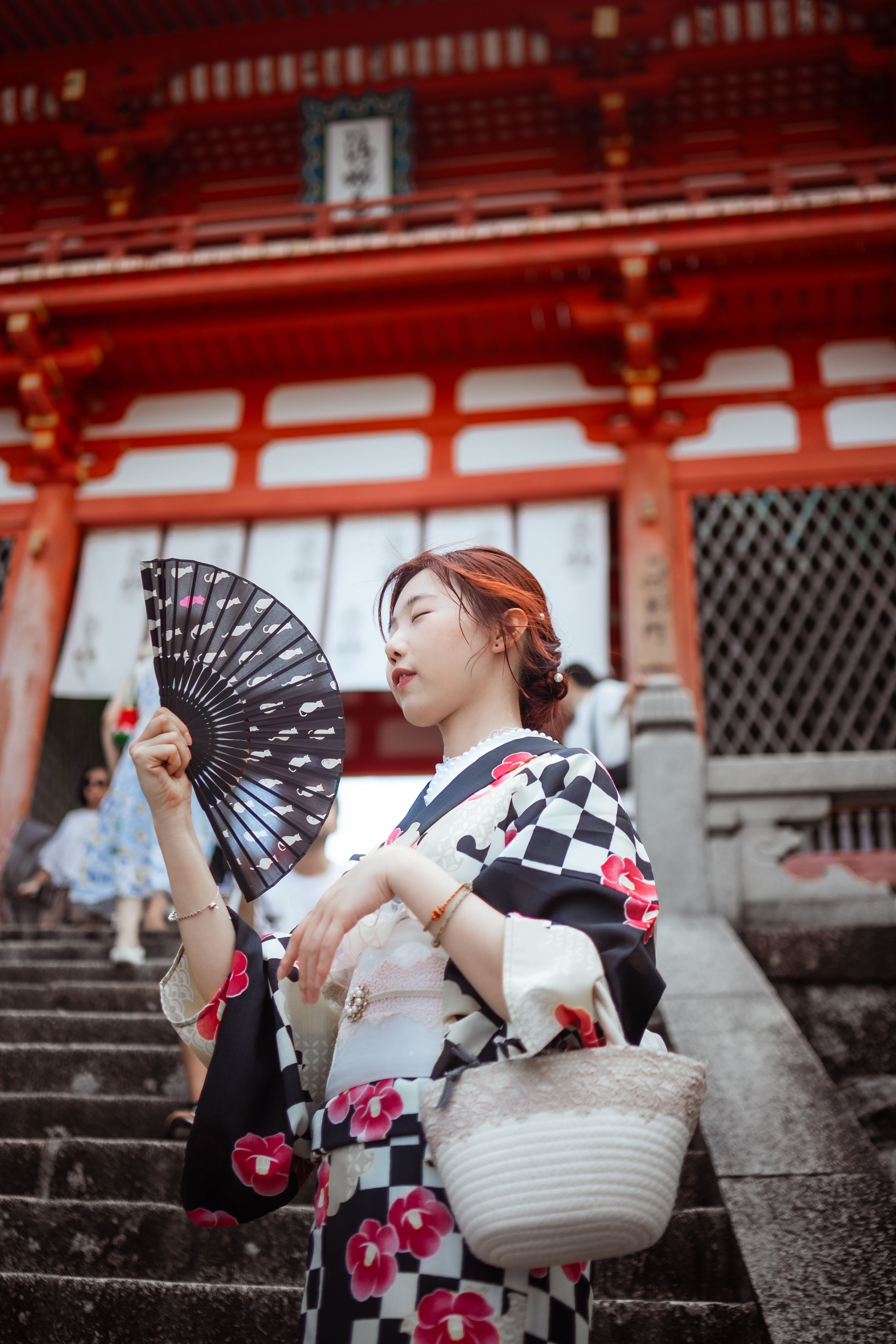 A young woman in a black-and-white checkered kimono with a pink floral pattern stands on stone steps, eyes closed, holding an open black fan beside her face, with the red beams and lattice windows of a Japanese shrine gate blurred behind her.