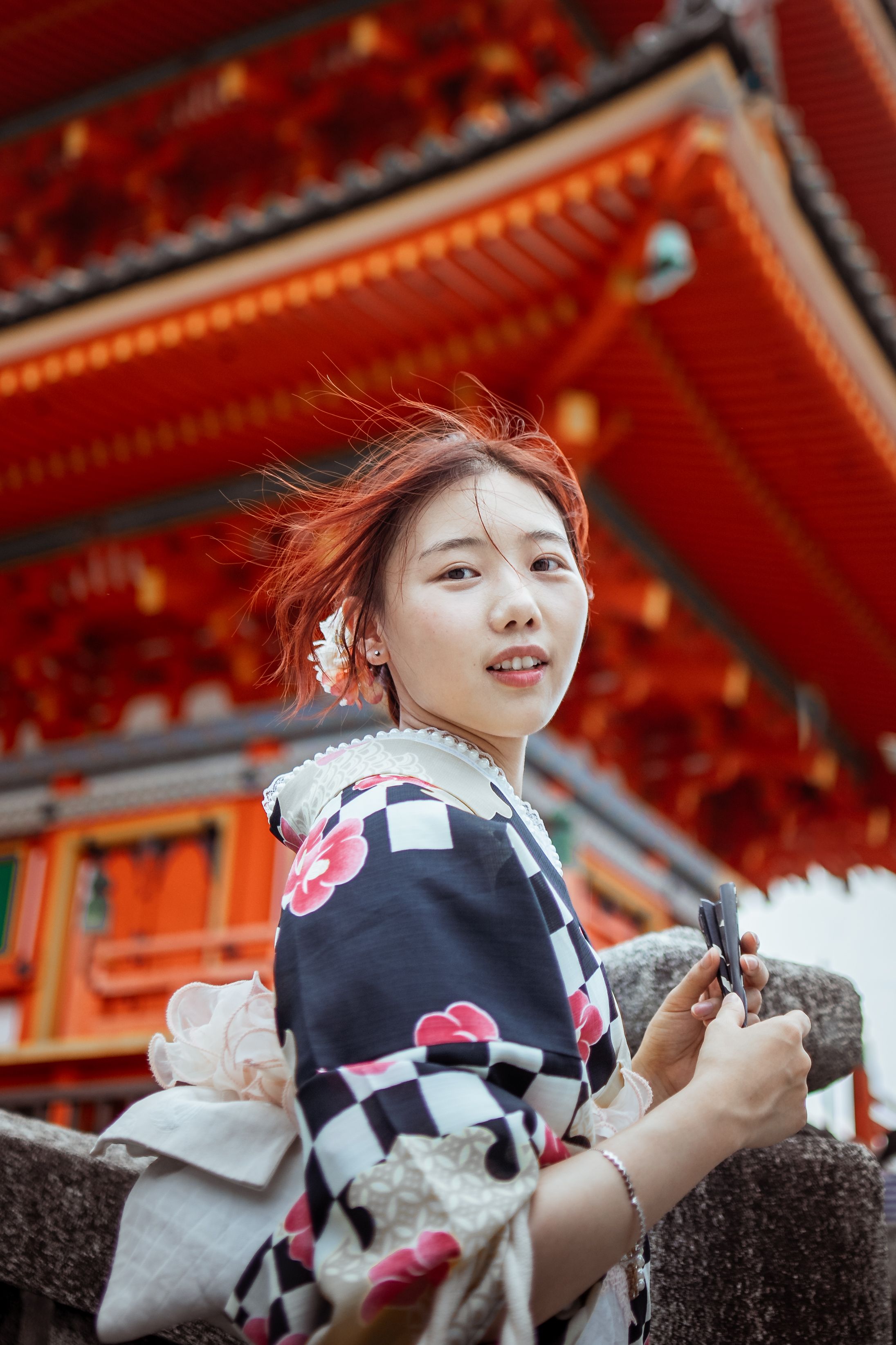 A young woman in a checkered kimono with pink floral patterns glances back over her shoulder, wind blowing through her hair, one hand holding a folding fan and resting on a stone railing, with the red wooden eaves of a temple roof looming behind her.
