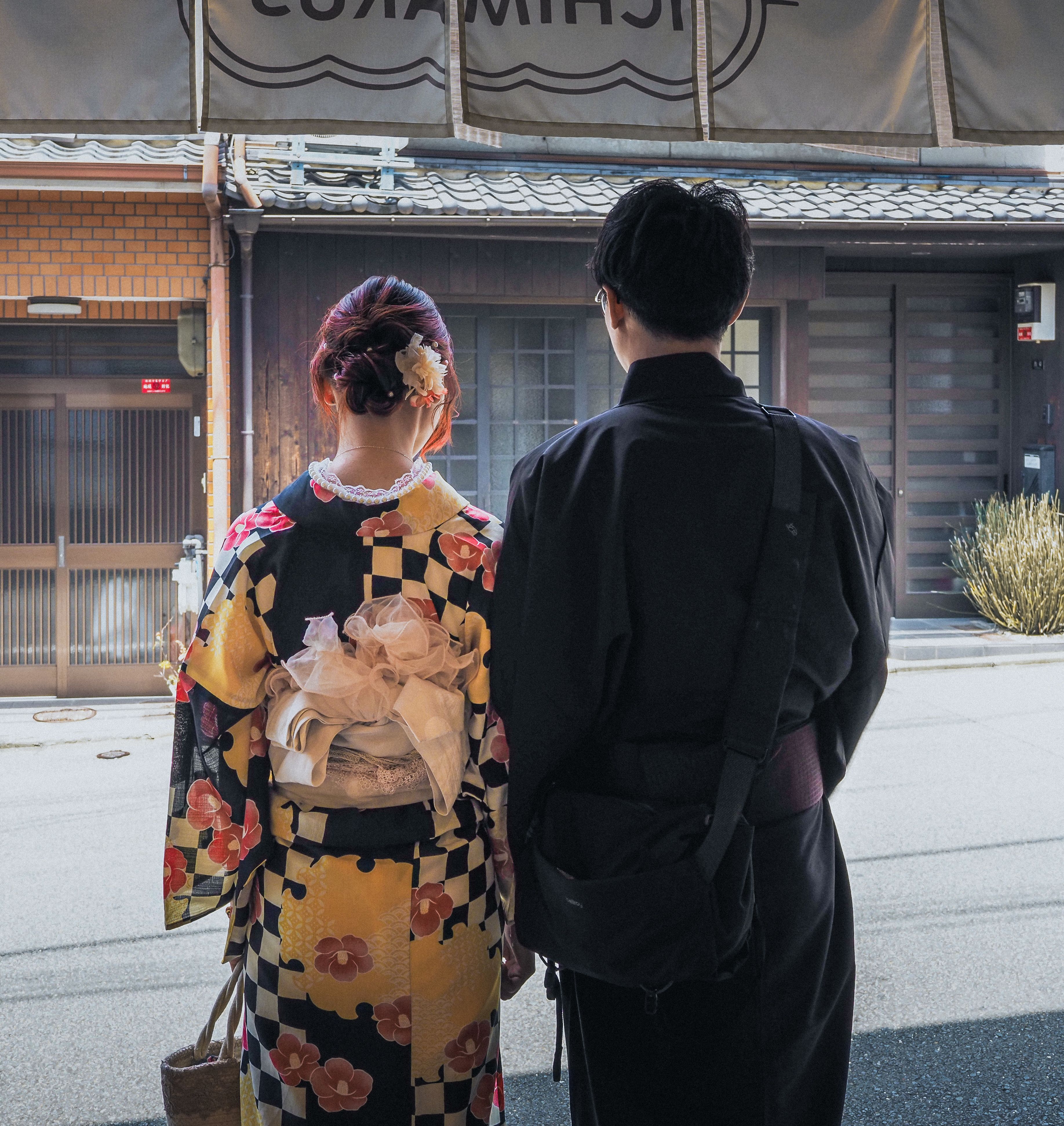 Two people are photographed from behind, standing side by side on a paved street in daylight; one has an upswept hairstyle with a white flower ornament and wears a black, gold, and pink floral kimono with a pale ruffled sash tied at the back, and the other wears a plain dark kimono, both facing a traditional wooden building with a tiled roof and a wide fabric banner hanging above its entrance.