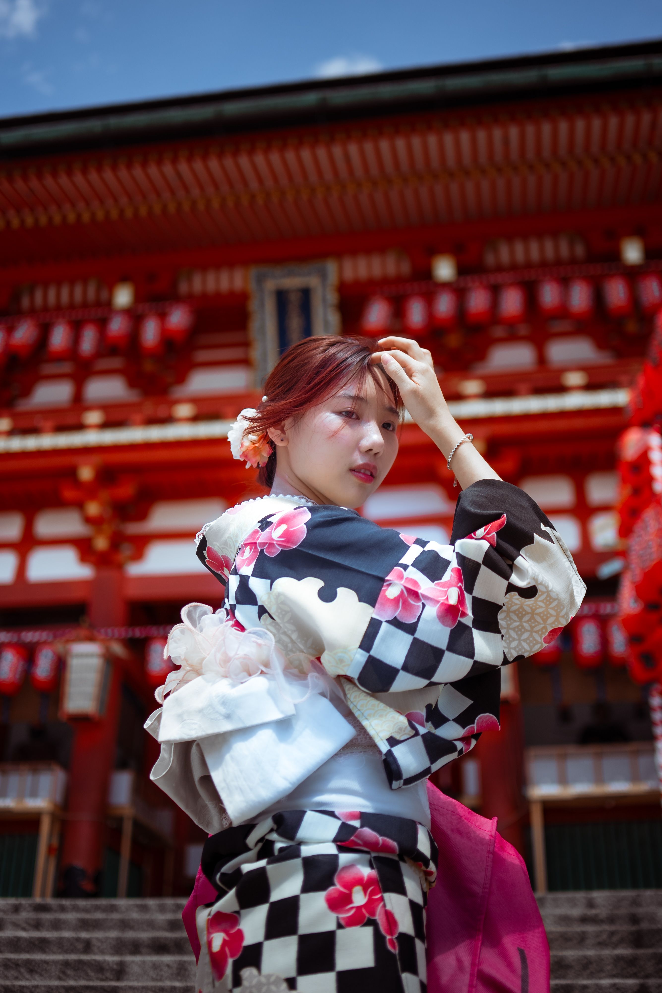 A woman in a black-and-white checkered kimono patterned with pink flowers stands on stone steps before a red Japanese shrine building strung with red lanterns, one hand raised to her hair as she glances back over her shoulder under a blue sky.