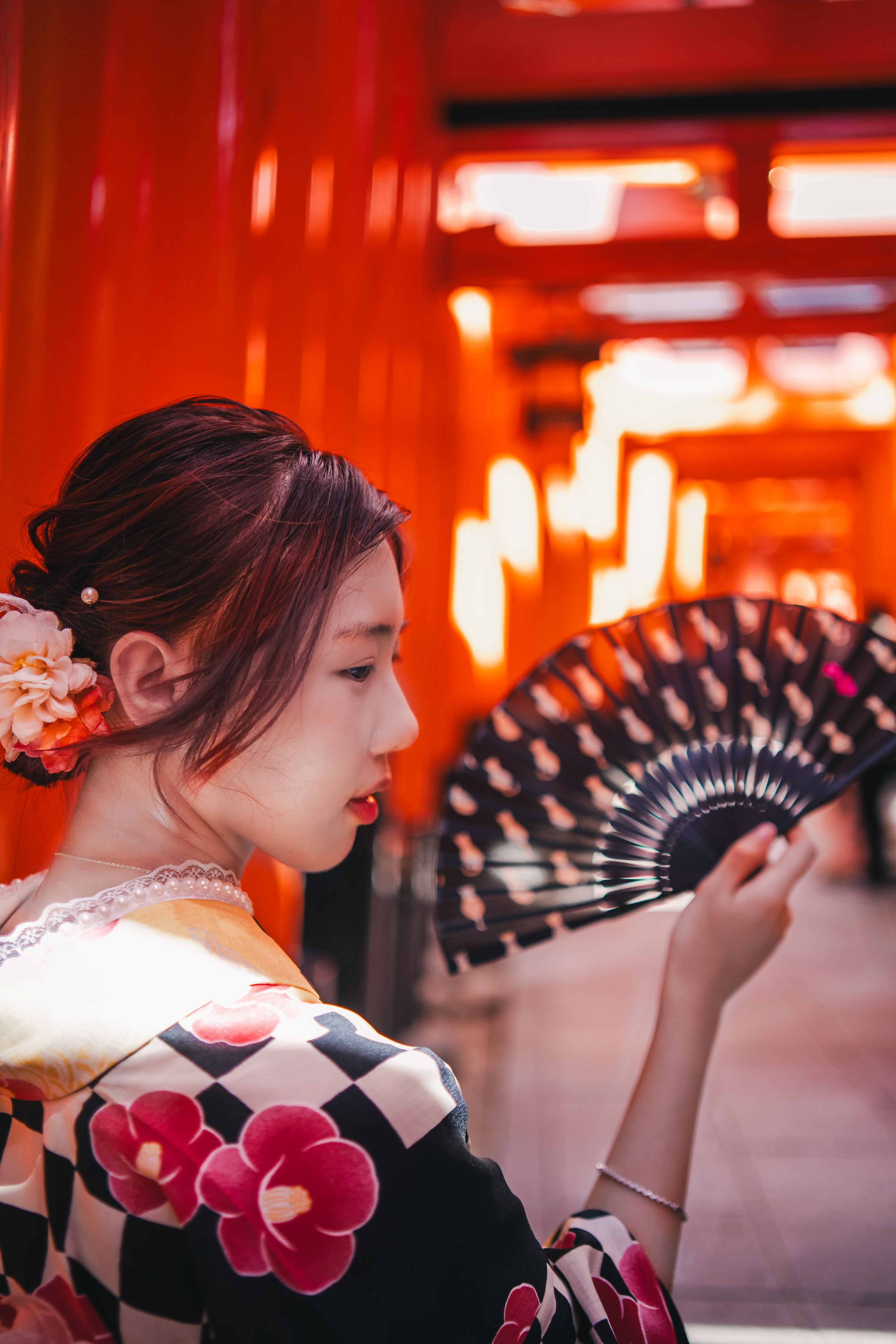 A woman in a floral kimono is photographed in profile, raising a dark patterned folding fan near her face, with a row of blurred red torii gate posts glowing softly behind her.