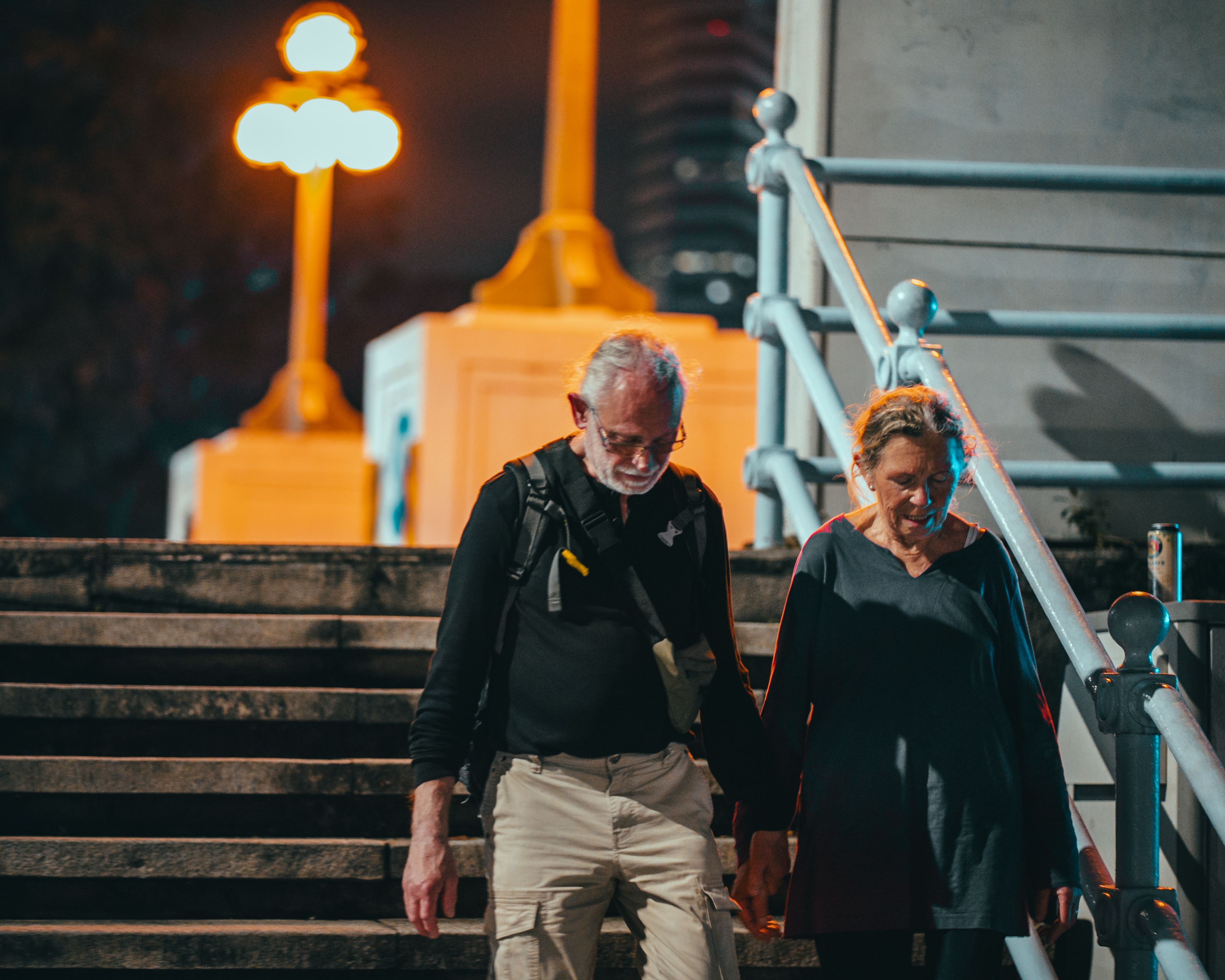 Two grey-haired adults, a man in a dark top and khaki cargo pants and a woman in a dark top, walk down an outdoor stone staircase at night holding hands, both looking down at their feet, with a glowing orange lamppost and a pale metal handrail in the background.