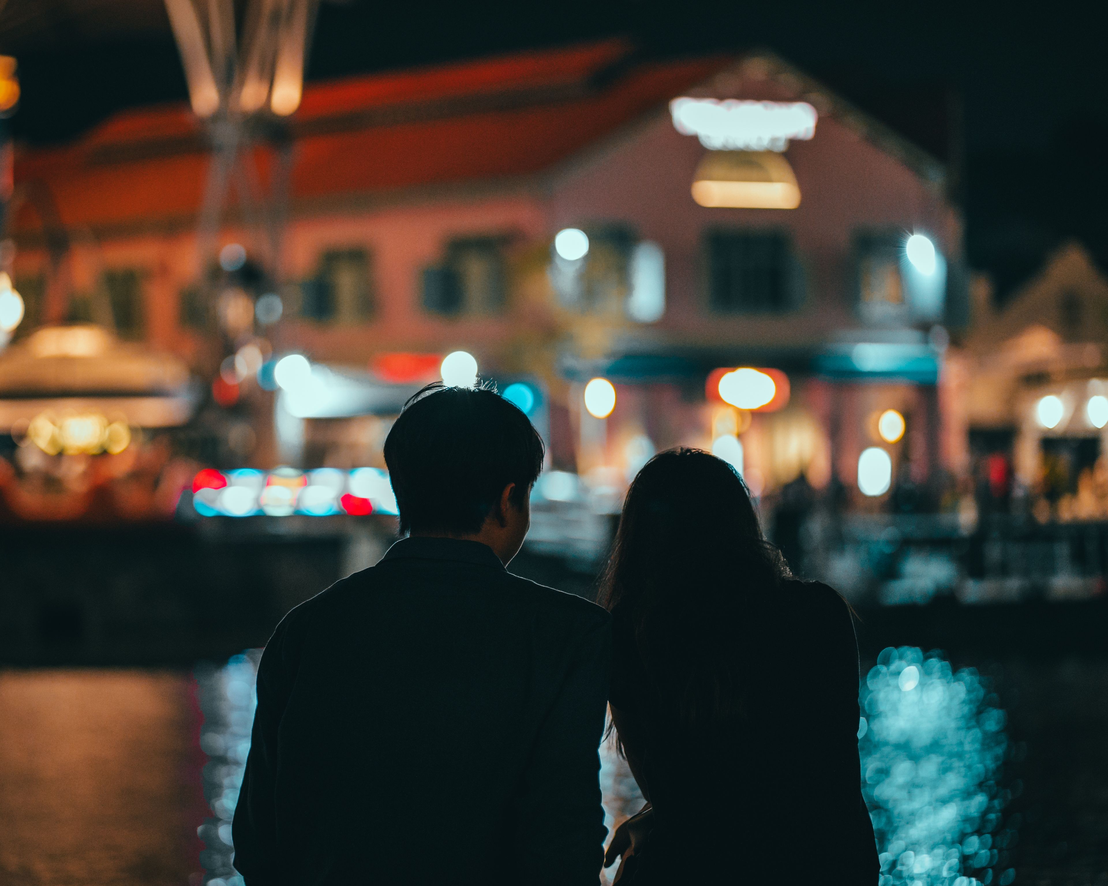 Two people are seen from behind in silhouette at night, standing close together and facing a waterfront row of illuminated buildings, with colorful lights blurred into soft bokeh across the foreground and background.