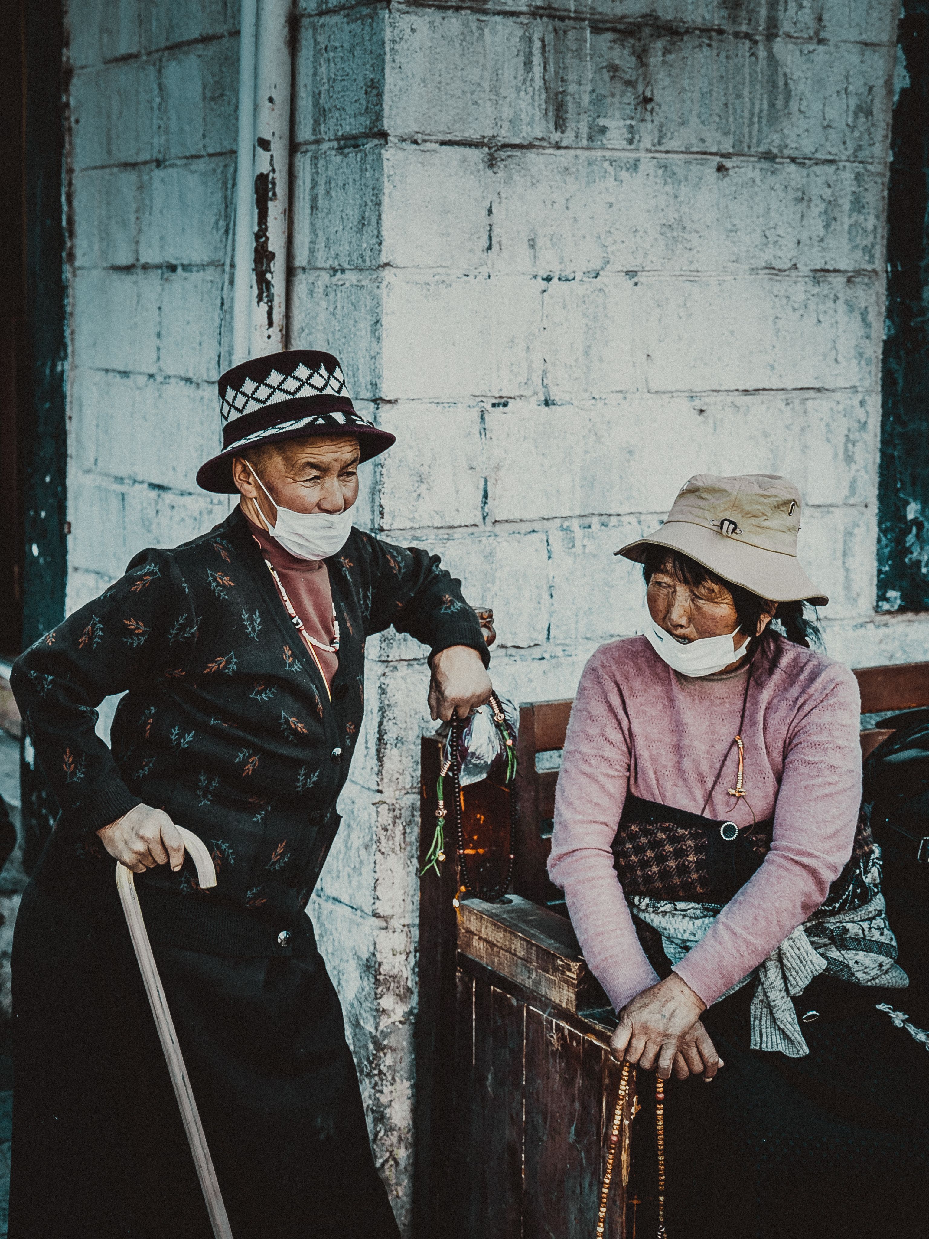 Two masked elderly people stand in daylight against a rough, whitewashed concrete block wall, one wearing a patterned hat and dark cardigan and leaning on a wooden cane, the other in a beige bucket hat and pink sweater leaning on a wooden railing, both holding strings of prayer beads.