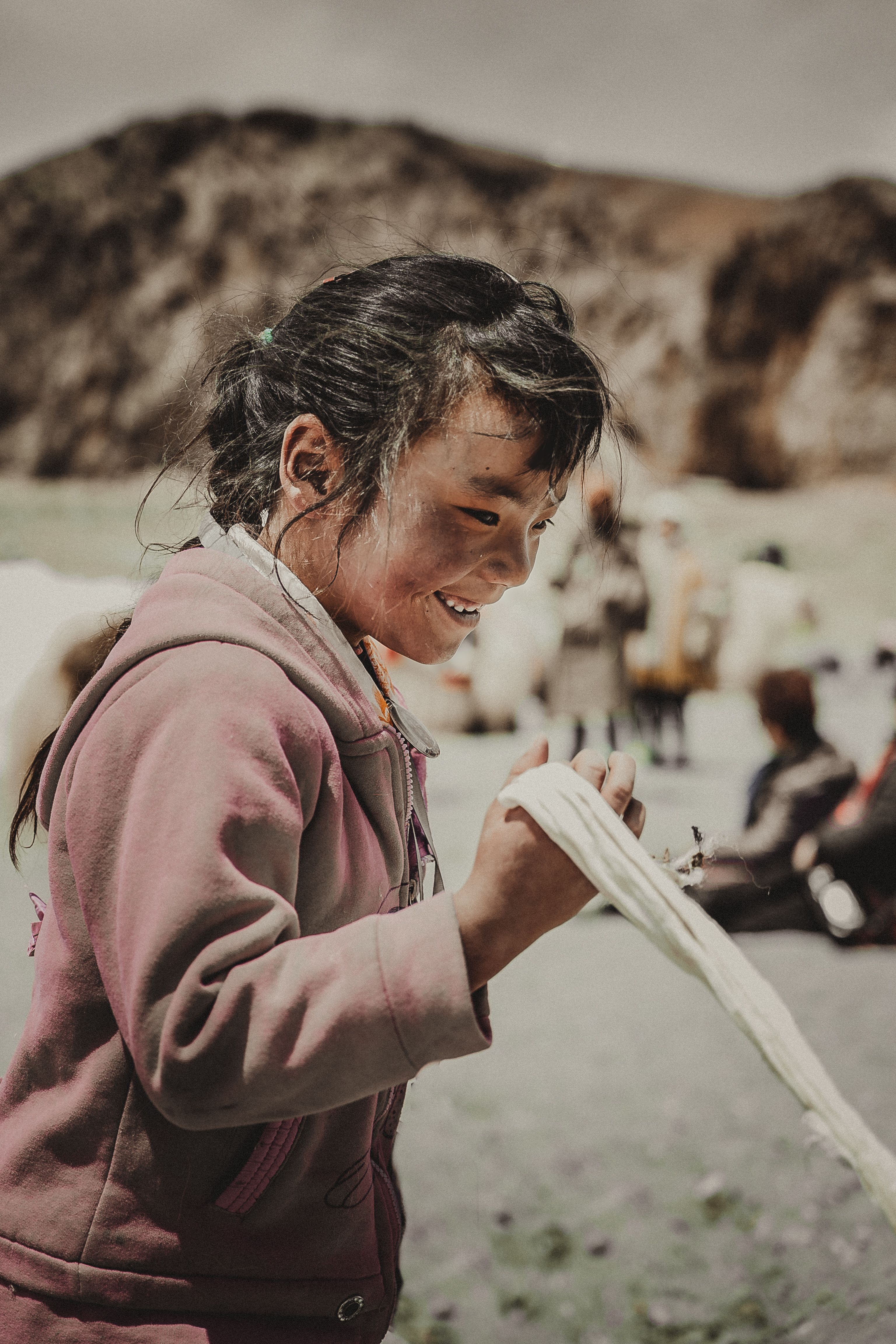 A child in a pink hoodie smiles while stretching a long strand of raw white wool between the fingers of both hands, with blurred figures in pale clothing and hazy hills in the background.