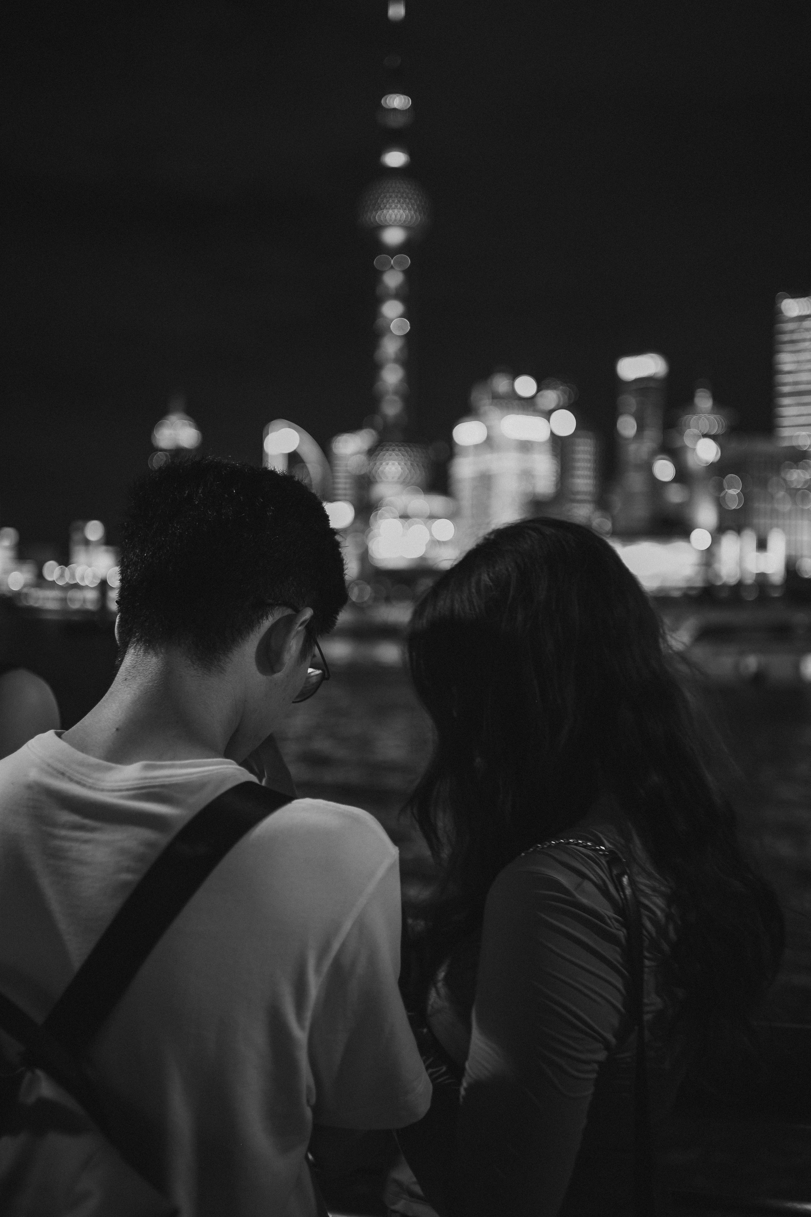 Black-and-white night photograph of two people seen from behind, one with short hair and glasses wearing a light T-shirt and the other with long dark hair, both facing a brightly lit city skyline with a tall illuminated tower blurred in the background.
