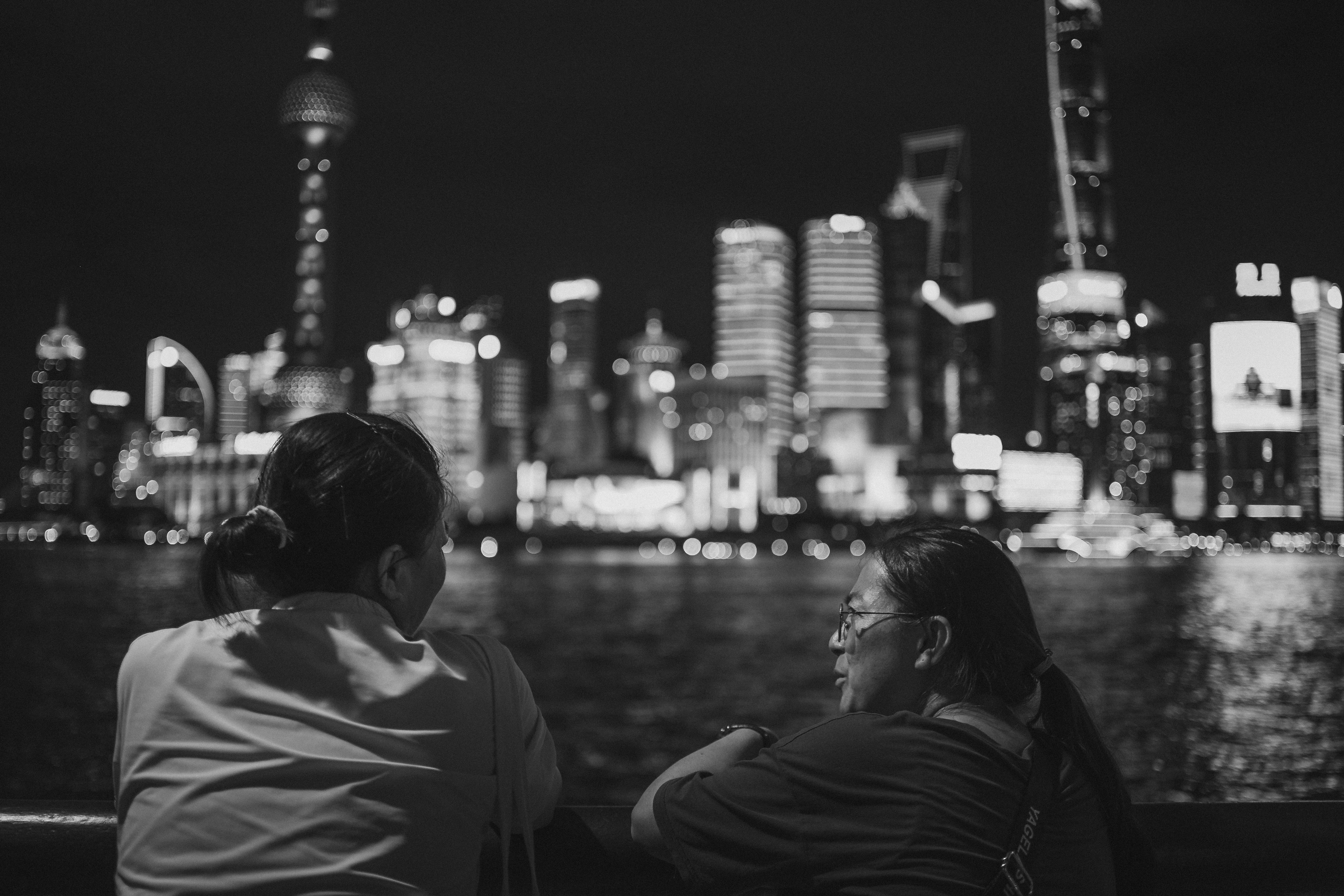 Black-and-white photograph of two people seen from behind and in profile, leaning on a railing as they look out across a river toward a brightly lit city skyline rendered in soft, blurred bokeh at night.
