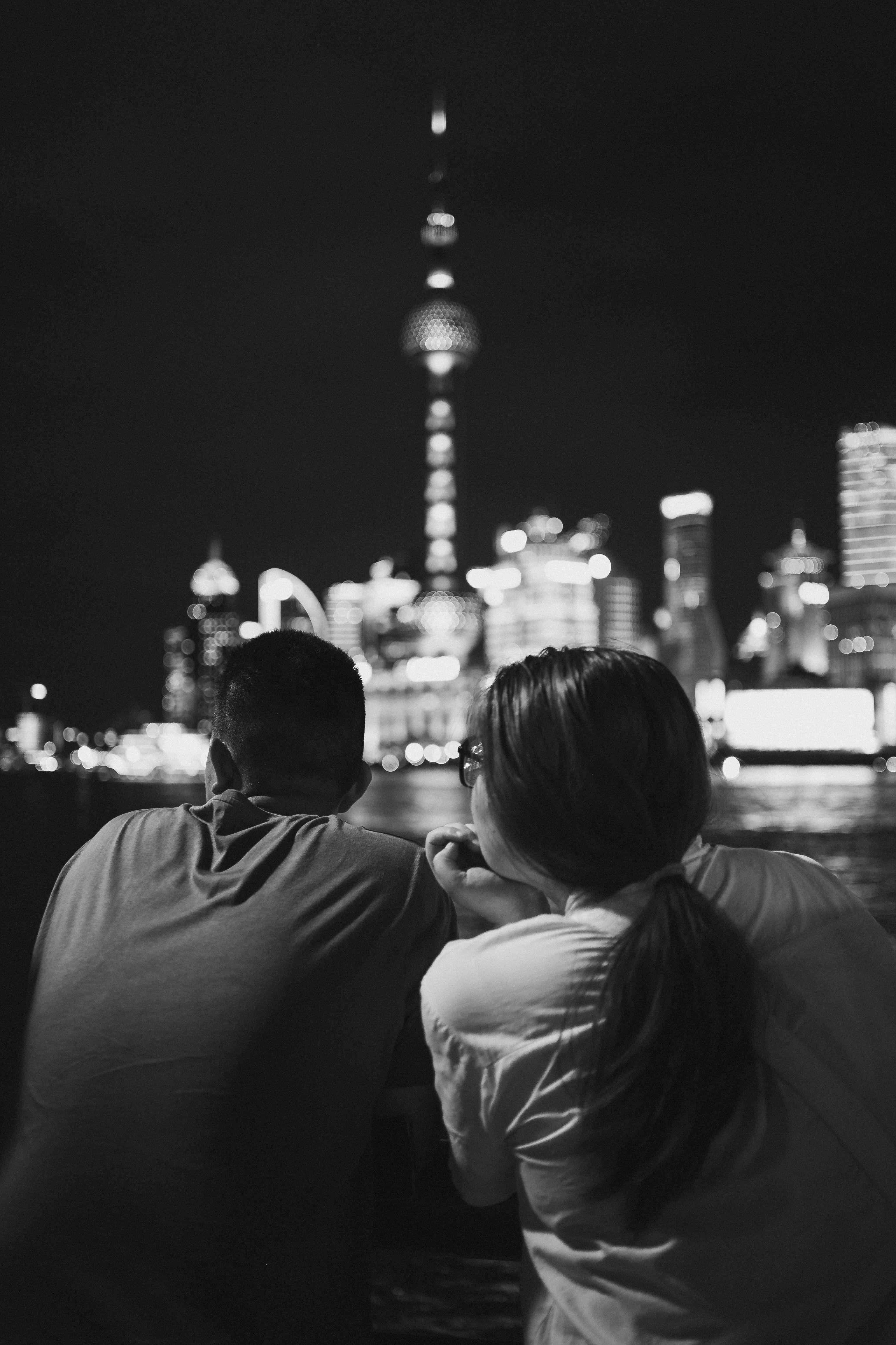 Black-and-white photo taken from behind of two people sitting close together, shoulders touching, facing a nighttime city skyline across water with a tall illuminated tower and softly blurred bokeh lights filling the background.