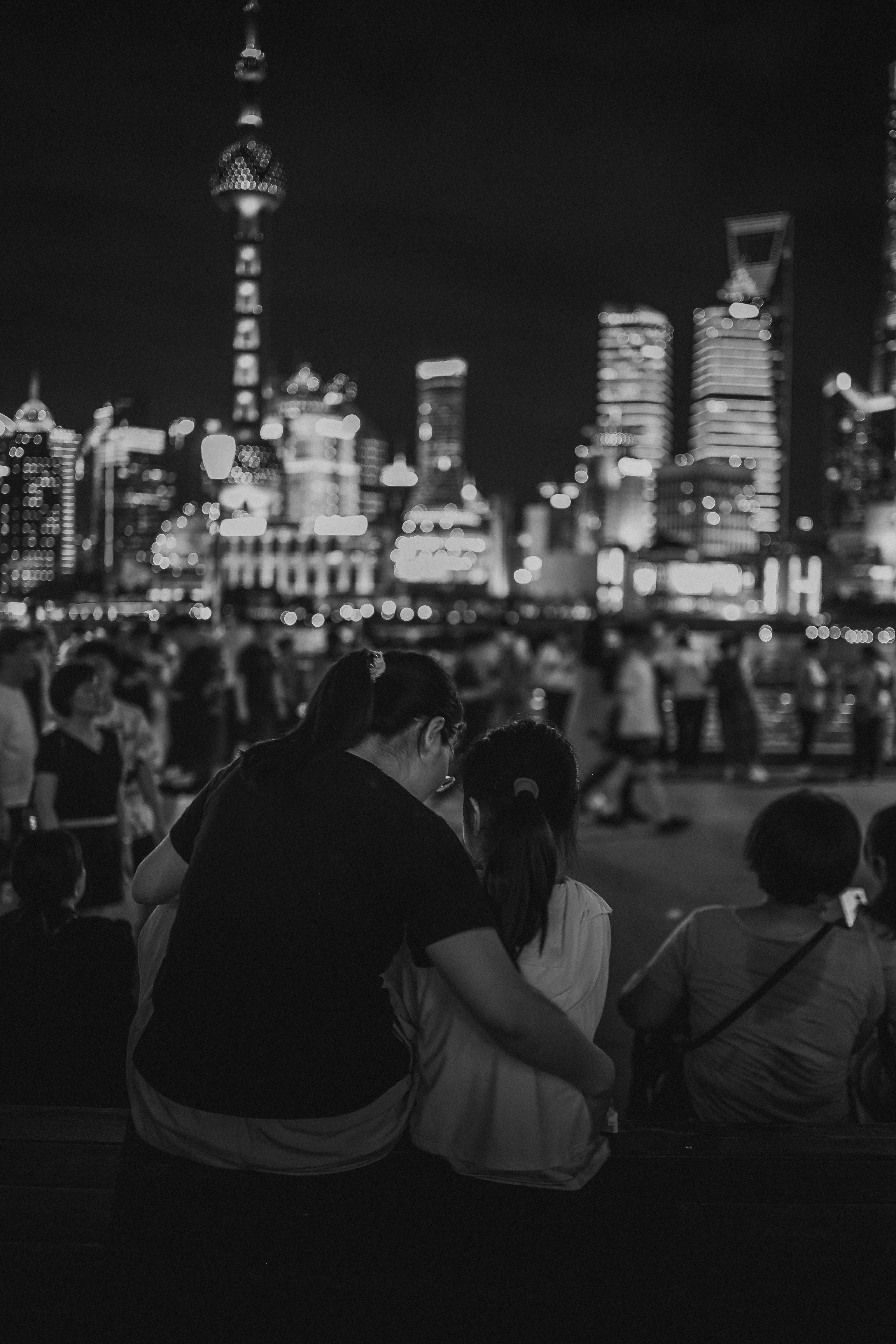 Black-and-white night photograph, shot from behind, of an adult with an arm around a child seated among other onlookers, all facing a brightly lit high-rise city skyline that dissolves into soft bokeh in the darkness.