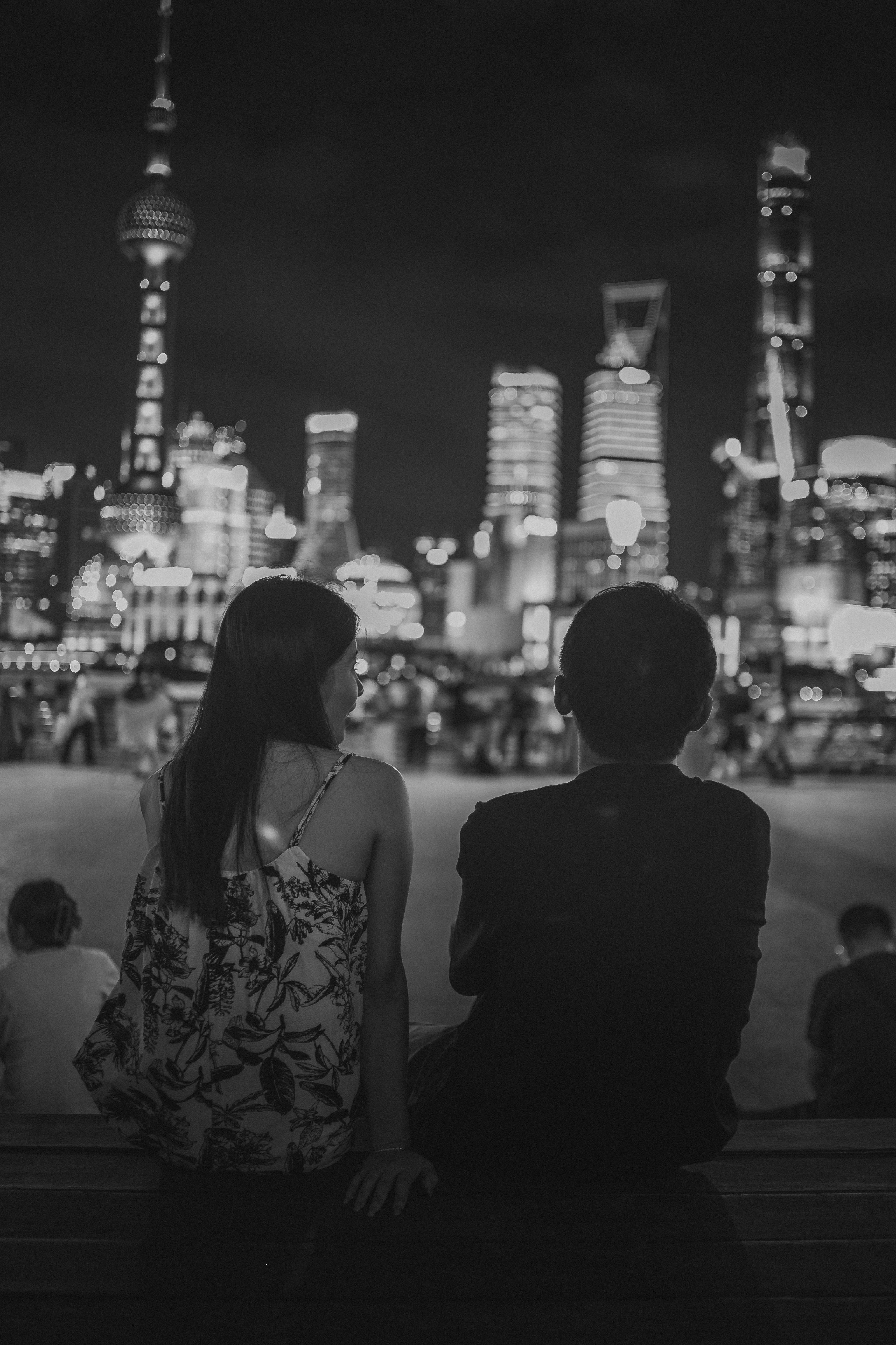 Black-and-white night photograph of two people seen from behind, sitting side by side in silhouette on a ledge, facing an illuminated city skyline with a spire-like tower topped by spheres and several skyscrapers, with other blurred figures seated nearby.