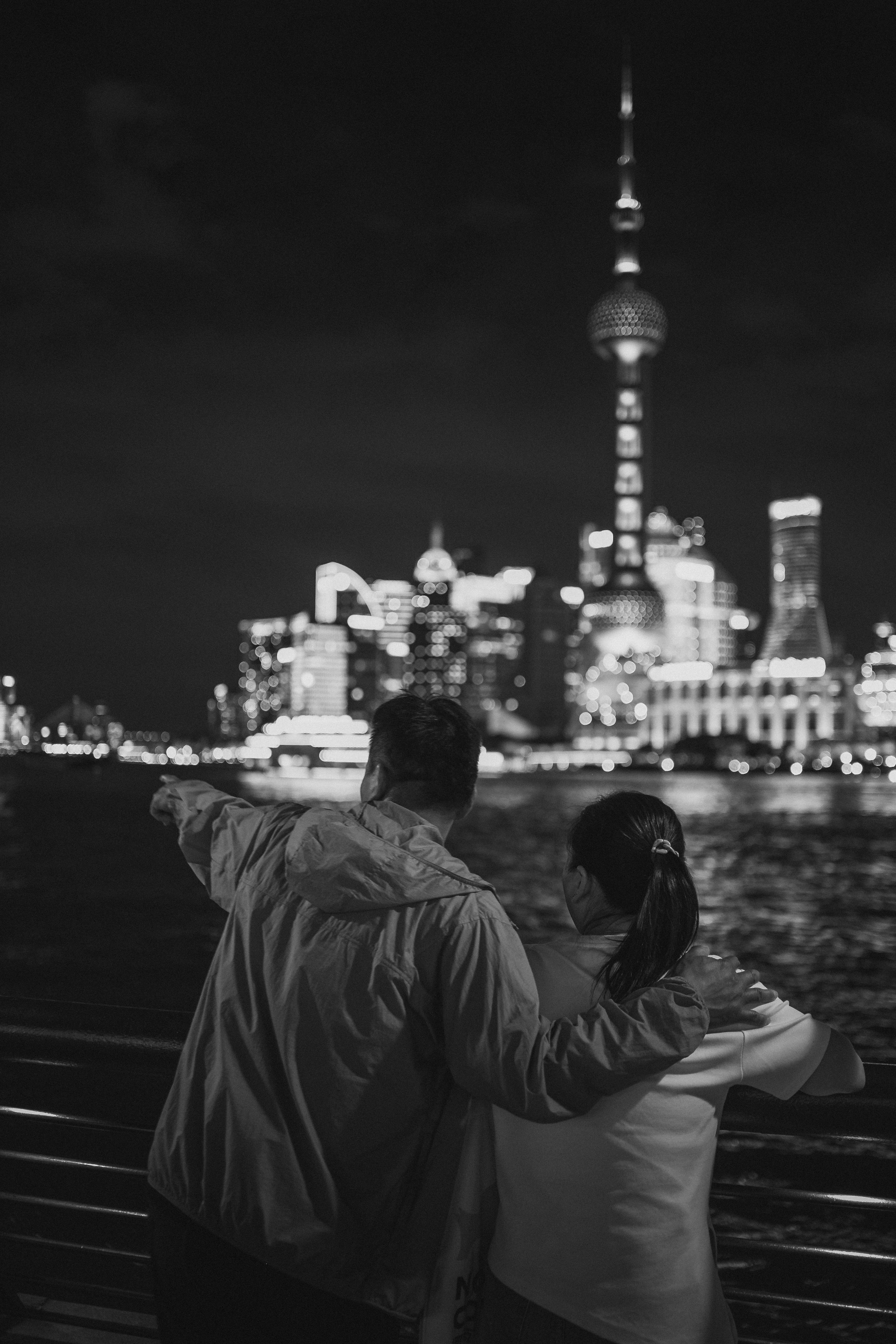 A black-and-white photograph shows two people from behind at a waterfront railing at night, one with an arm raised and pointing toward a brightly lit, out-of-focus city skyline topped by a tall spired tower across the water.