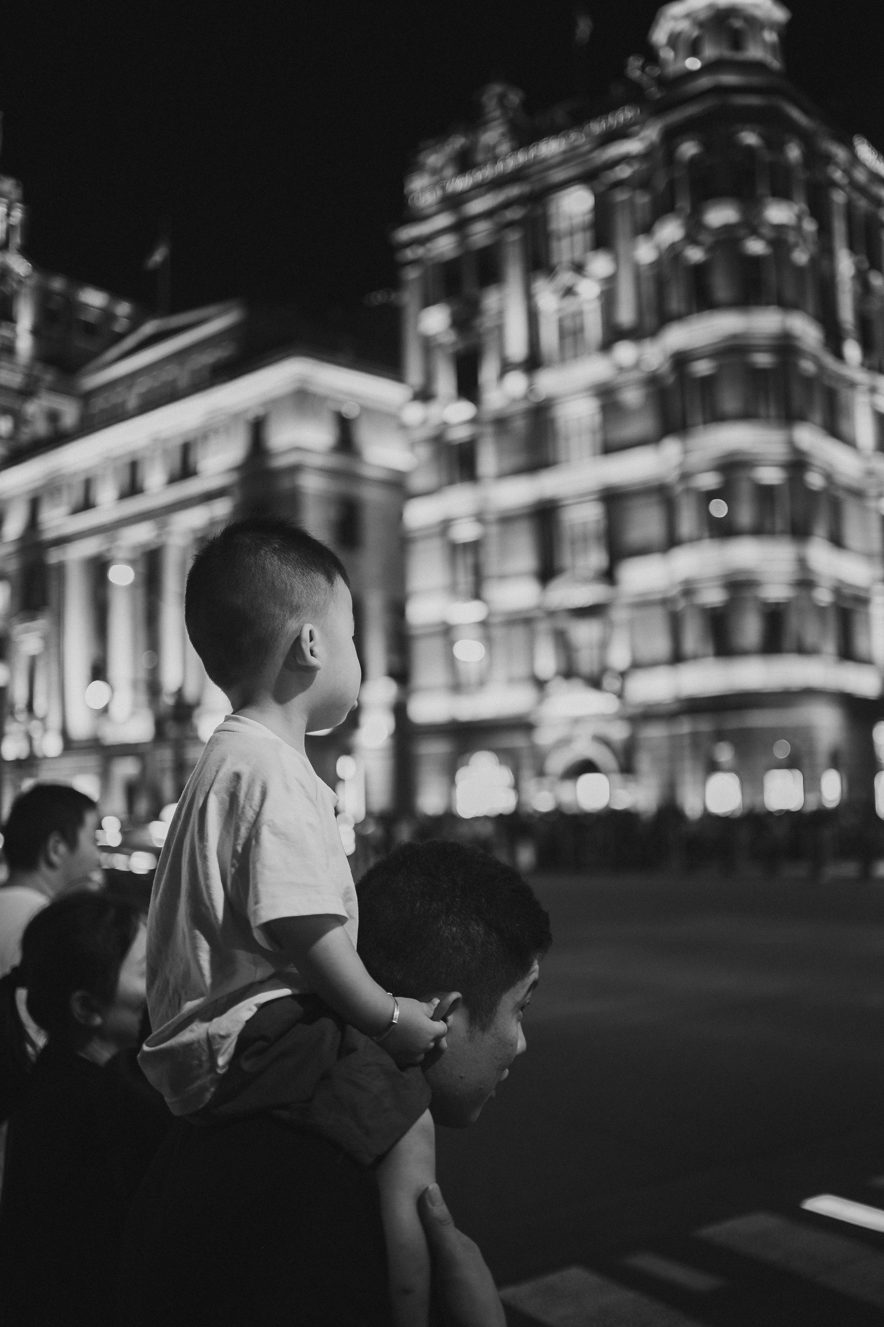 Black-and-white night photograph of a young child sitting on an adult's shoulders and holding the adult's hands, both seen from the side facing away toward a row of illuminated, ornately detailed buildings, with a blurred figure of another person visible at the lower left.